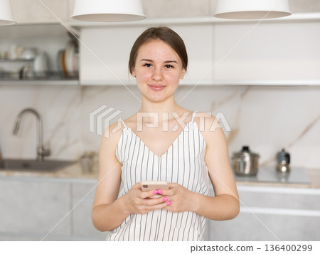 Young girl standing in the kitchen holding mobile phone Young girl standing in the kitchen holding mobile phone 136400299