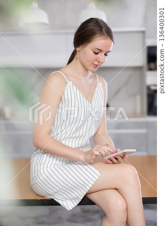In kitchen, positive girl in casual clothes sits on table and chatting in smartphone In kitchen, positive girl in casual clothes sits on table and chatting in smartphone 136400301
