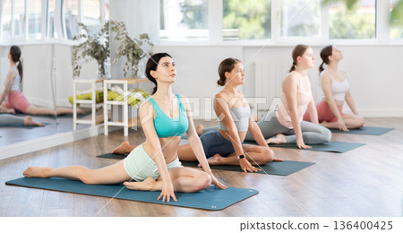 Group of women practicing pigeon pose of yoga on black mat in fitness studio 136400425