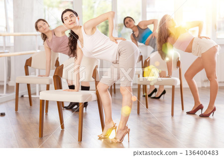 Leaning on chair seat with hand foot, young woman performs lap dance during group workout. Leaning on chair seat with hand foot, young woman performs lap dance during group workout. 136400483