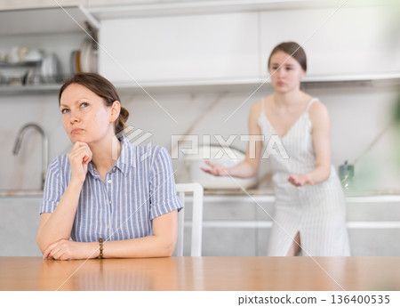 Offended mother sitting at the kitchen table, daughter standing behind Offended mother sitting at the kitchen table, daughter standing behind 136400535
