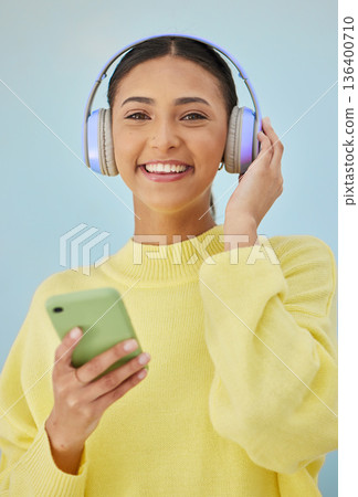 Portrait of woman with phone, headphones and smile in studio for social media, mobile app and streaming radio site. Podcast, music media subscription and happy girl with cellphone on blue background. Portrait of woman with phone, headphones and smile in studio for social media, mobile app and streaming radio site. Podcast, music media subscription and happy girl with cellphone on blue background. 136400710