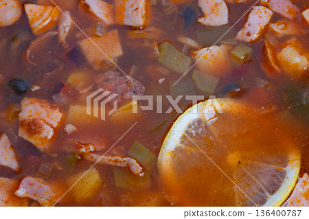 Rich tomato solyanka with lemon slices and diced smoked meats, olives, potato cubes in glossy aromatic broth, closeup texture shot conveying cozy home cooking and winter comfort 136400787
