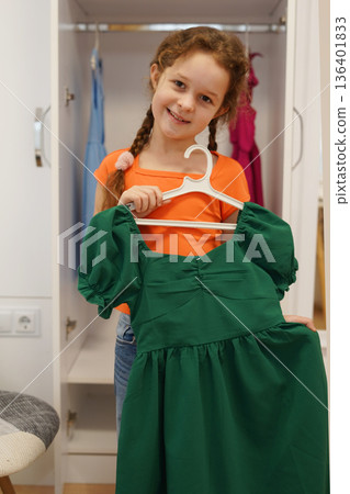Happy child girl holding green dress in white wardrobe smiling 136401833