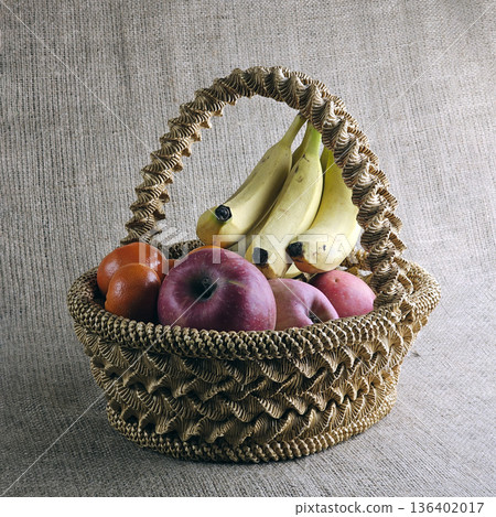 Woven straw basket against burlap background. Close-up. Eco-friendly. The handmade basket is filled with fruit Woven straw basket against burlap background. Close-up. Eco-friendly. The handmade basket is filled with fruit 136402017