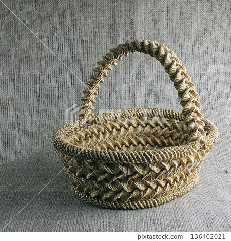 A woven straw basket against a burlap background. Close-up. Eco-friendly. Handmade basket for interior decoration and use as container for collecting fruits, vegetables, and mushrooms. A woven straw basket against a burlap background. Close-up. Eco-friendly. Handmade basket for interior decoration and use as container for collecting fruits, vegetables, and mushrooms. 136402021