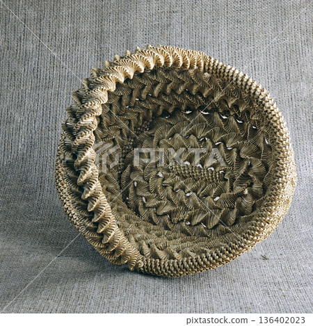Woven straw basket against a burlap background. Close-up. Eco-friendly. Handmade basket for interior decoration and use as container for collecting fruits, vegetables, and mushrooms. Woven straw basket against a burlap background. Close-up. Eco-friendly. Handmade basket for interior decoration and use as container for collecting fruits, vegetables, and mushrooms. 136402023