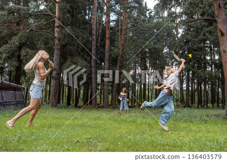 Active family enjoying outdoor leisure together, playing badminton on grass meadow with pine trees. Mother and children having fun in nature, summer weekend, wide angle full length Active family enjoying outdoor leisure together, playing badminton on grass meadow with pine trees. Mother and children having fun in nature, summer weekend, wide angle full length 136403579