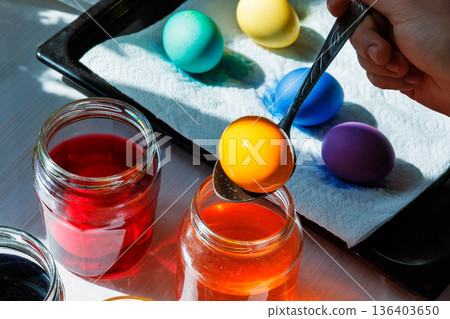 Close up of the spoon lifting orange dyed egg above jar with colorful easter eggs on tray 136403650