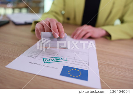 Hand placing an approval stamp on an official document with a European Union logo at a bright modern office 136404004