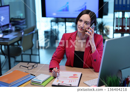 Business professional in a modern office engaged in a phone call while analyzing charts and data reports 136404218