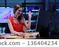 Professional woman engaging in a virtual meeting while smiling and waving in a modern office setting during evening hours 136404234