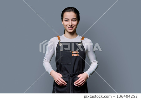 Portrait of young smiling female student in apron on gray background Portrait of young smiling female student in apron on gray background 136404252