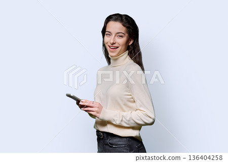 Portrait of smiling teenage girl holding smartphone on white studio background 136404258