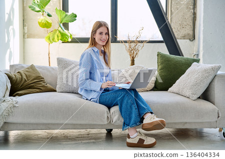 Portrait of young woman sitting on sofa typing on laptop, in home interior 136404334