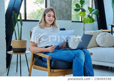 Young smiling student girl sitting at home on chair with laptop, reading papers Young smiling student girl sitting at home on chair with laptop, reading papers 136404340