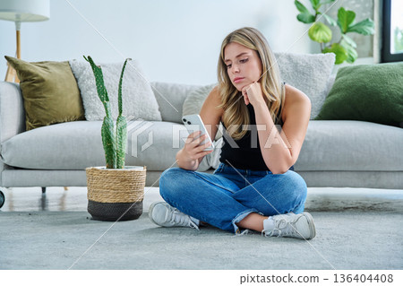 Serious young woman with smartphone sitting at home near sofa 136404408