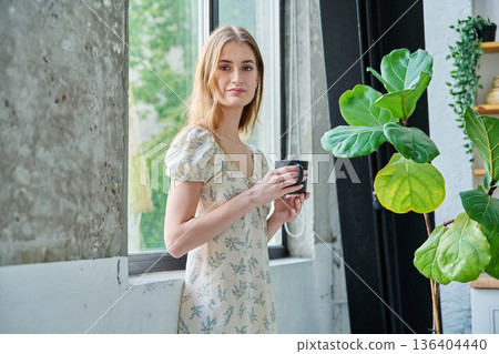 Young beautiful smiling romantic girl looking at camera with cup, standing near window Young beautiful smiling romantic girl looking at camera with cup, standing near window 136404440