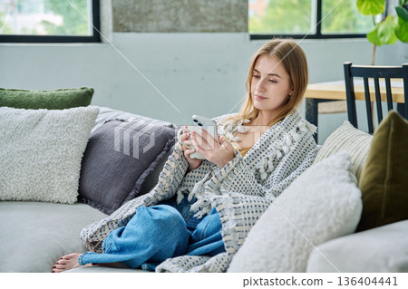 Relaxed beautiful young woman sitting at home on sofa with cup, using smartphone under blanket Relaxed beautiful young woman sitting at home on sofa with cup, using smartphone under blanket 136404441