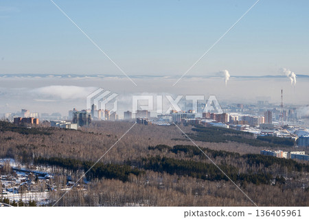 Winter cityscape of Krasnoyarsk with urban buildings and an industrial pipe emitting smoke into the polluted sky, showing environmental concern. 136405961