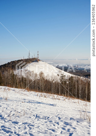 Winter landscape of Nikolaevskaya Sopka, Gremyachaya Griva park in Krasnoyarsk, Russia. Snow-covered hill with communication towers and a city view on a clear day 136405962