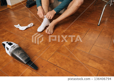 Close-up cropped shot of man sitting on wooden floor removing white sock, holding leg, with a handheld vacuum cleaner and upright vacuum in background, representing domestic routine and self-care. Close-up cropped shot of man sitting on wooden floor removing white sock, holding leg, with a handheld vacuum cleaner and upright vacuum in background, representing domestic routine and self-care. 136406794