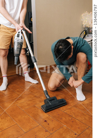 Vertical shot of two men performing household maintenance together, with one using vacuum to tidy up wooden flooring, another prepares for activity, emphasizing teamwork and domestic responsibility. Vertical shot of two men performing household maintenance together, with one using vacuum to tidy up wooden flooring, another prepares for activity, emphasizing teamwork and domestic responsibility. 136406797