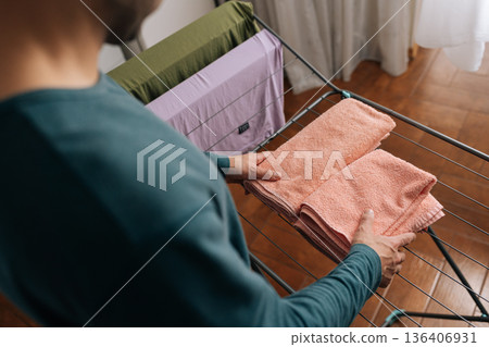 View from shoulder of unrecognizable person folding clean, soft pink towels on laundry drying rack, performing daily household chores and maintaining fresh, organized linens indoors. 136406931