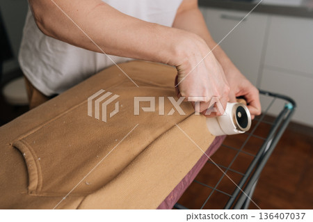 Male hands using battery lint remover on brown sweater, shaving away fuzz and pilling to restore smooth texture and refresh worn knitwear for everyday clothing care. Concept of effective fabric care. Male hands using battery lint remover on brown sweater, shaving away fuzz and pilling to restore smooth texture and refresh worn knitwear for everyday clothing care. Concept of effective fabric care. 136407037