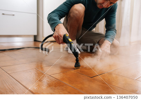 Focused man steaming and scrubbing herringbone wood-look tile floor with brush attachment, removing grime and sanitizing grout for a fresh, spotless, healthy home environment. Focused man steaming and scrubbing herringbone wood-look tile floor with brush attachment, removing grime and sanitizing grout for a fresh, spotless, healthy home environment. 136407050
