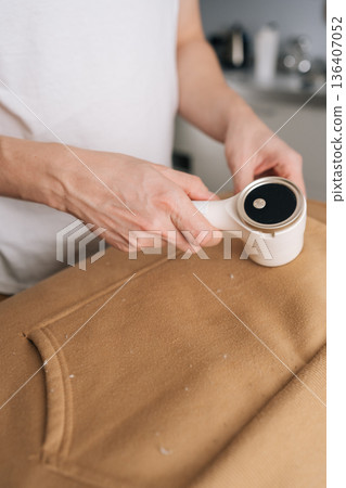 Vertical cropped shot of person using electric fabric shaver, removing pilling and fuzz from sweatshirt, maintaining clothing appearance and textile quality through effective garment care, close-up. Vertical cropped shot of person using electric fabric shaver, removing pilling and fuzz from sweatshirt, maintaining clothing appearance and textile quality through effective garment care, close-up. 136407052