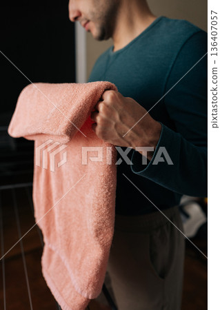 Close-up vertical cropped shot of man standing in home, holding clean, dry, folded pink towel, reflecting modern domesticity, shared household responsibilities and gender equality in chores. Close-up vertical cropped shot of man standing in home, holding clean, dry, folded pink towel, reflecting modern domesticity, shared household responsibilities and gender equality in chores. 136407057