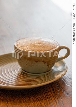A warm ceramic coffee cup with foam sits on a matching saucer atop a wooden table 136407190