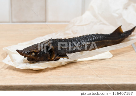 A whole hot smoked sterlet fish lies on crumpled parchment atop a wooden table 136407197