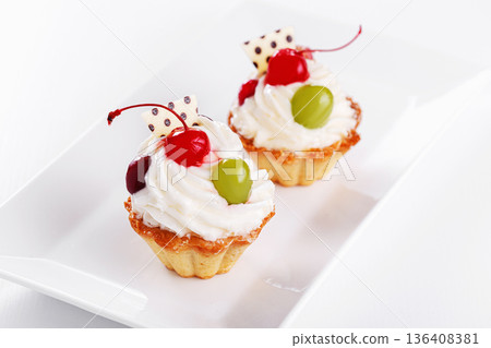 Beautiful cream cakes with cherries and grapes on a plate on a white background Beautiful cream cakes with cherries and grapes on a plate on a white background 136408381