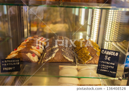 Traditional Italian cannoli pastries with various toppings displayed in a bakery glass showcase, featuring classic Sicilian desserts under warm interior lighting. 136408526