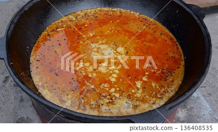 Big pot simmers, Iron skillet filled with fragrant stew bubbling outside, Large vessel containing savory liquid cooking over open air, Spiced liquid stew gently boiling inside sturdy iron pan outdoors Big pot simmers, Iron skillet filled with fragrant stew bubbling outside, Large vessel containing savory liquid cooking over open air, Spiced liquid stew gently boiling inside sturdy iron pan outdoors 136408535