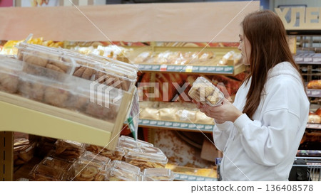 Young female shopper browsing the bakery section of a supermarket, holding a container of cookies and exploring the assortment on offer Young female shopper browsing the bakery section of a supermarket, holding a container of cookies and exploring the assortment on offer 136408578