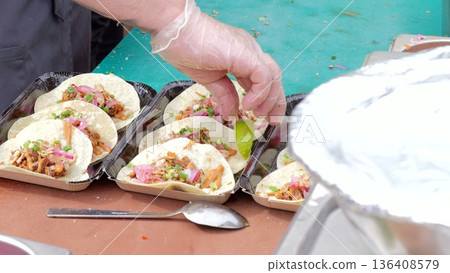 Tacos garnished with lime, Fingers placing lime on warm tacos display, Aromas and toppings on freshly prepared Mexican dishes, Serving counter scene with disposable plates and zesty taco Tacos garnished with lime, Fingers placing lime on warm tacos display, Aromas and toppings on freshly prepared Mexican dishes, Serving counter scene with disposable plates and zesty taco 136408579