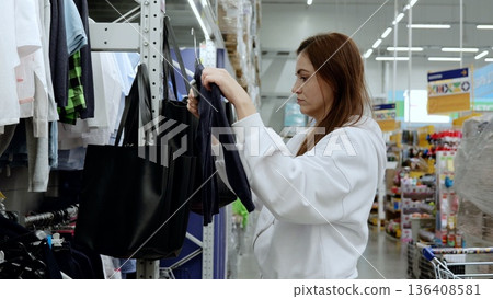 Woman enjoys a modern shopping experience, examining a blue garment in a bustling store with a black tote bag Woman enjoys a modern shopping experience, examining a blue garment in a bustling store with a black tote bag 136408581