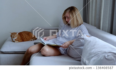 Tranquil domestic scene, woman reading with her ginger cat peacefully sleeping beside her on a comfortable sofa in a cozy living room Tranquil domestic scene, woman reading with her ginger cat peacefully sleeping beside her on a comfortable sofa in a cozy living room 136408587