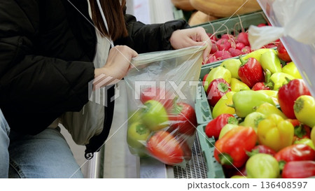 Customer filling plastic bag with fresh bell peppers at grocery store, selecting healthy ingredients for meal preparation 136408597
