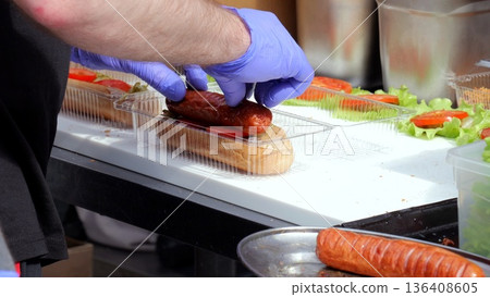 Food vendor preparing, Vendor assembling sausages at food festival, Closeup of hot dog preparation at lively outdoor event, Chef with gloves preparing sausage sandwiches amidst bustling food market Food vendor preparing, Vendor assembling sausages at food festival, Closeup of hot dog preparation at lively outdoor event, Chef with gloves preparing sausage sandwiches amidst bustling food market 136408605