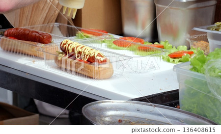 Gloved hands adjusting toppings on assembled frankfurter, vendor inspecting quality before serving, careful hygiene and final plating at busy outdoor market, honest preparation and customer service 136408615