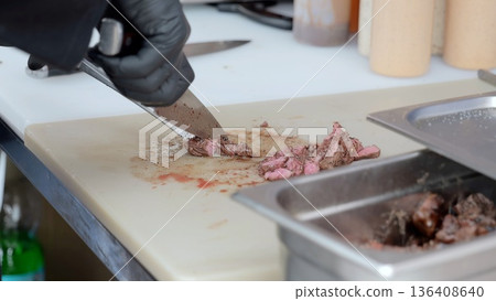 Hands slicing smoked brisket on board, vendor wearing black gloves carves tender brisket into thin slices on yellow board beside stainless tray, smoky aroma, casual outdoor festival catering scene Hands slicing smoked brisket on board, vendor wearing black gloves carves tender brisket into thin slices on yellow board beside stainless tray, smoky aroma, casual outdoor festival catering scene 136408640