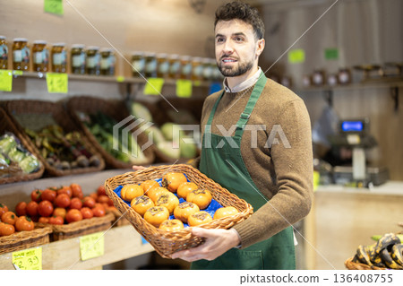 Young male seller holding basket of persimmons in grocery market 136408755