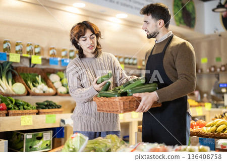 Girl customer in vegetable store examines zucchini, male seller advises and help with choice Girl customer in vegetable store examines zucchini, male seller advises and help with choice 136408758