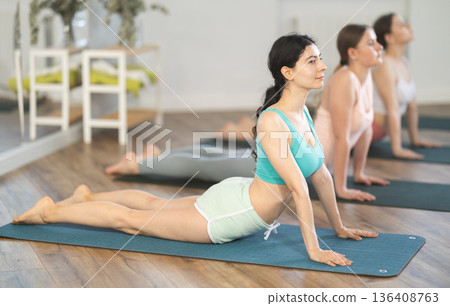 Women bending in back while lying on stomach in Bhujangasana Cobra Stretch position during training together in studio Women bending in back while lying on stomach in Bhujangasana Cobra Stretch position during training together in studio 136408763