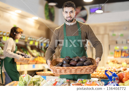 Young male seller holding basket of avocados standing by counter in grocery market 136408772