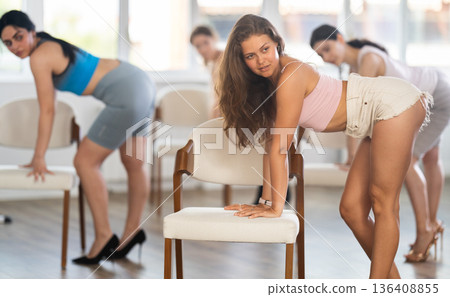 Leaning on chair seat with hand foot, young woman performs lap dance during group workout. Leaning on chair seat with hand foot, young woman performs lap dance during group workout. 136408855